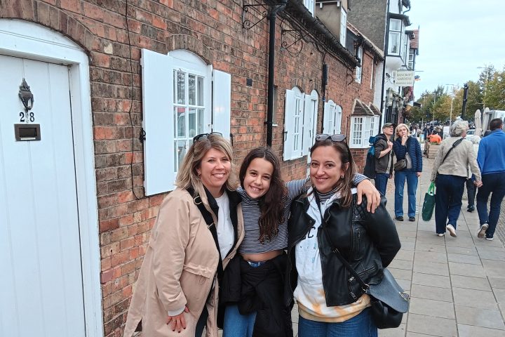 Three women smiling, standing on a street with brick buildings and passersby.