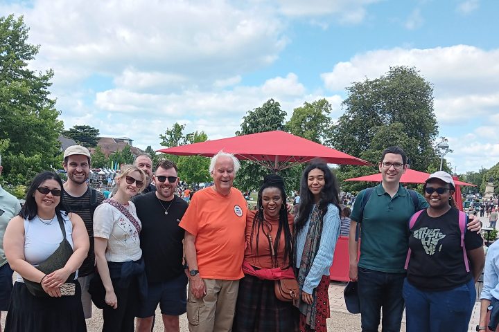 Group of people posing outdoors on a sunny day, with trees and a large red parasol in the background.