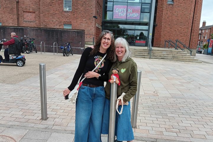 Two people with denim skirts smiling outside a building with a 'Gifts in the Shop' sign.
