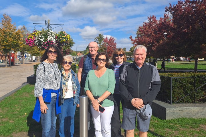 Group of six adults posing outdoors with trees and flowers in the background.