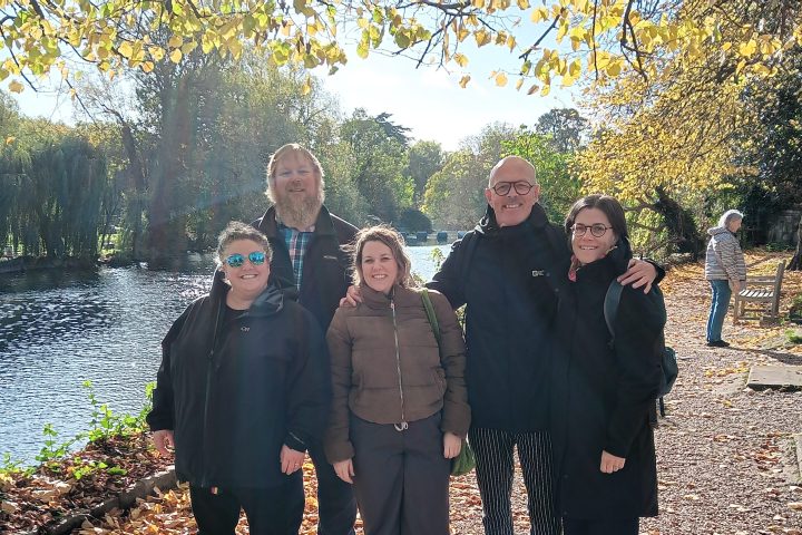 Five people smiling by a riverside with autumn leaves on the ground and trees in the background.
