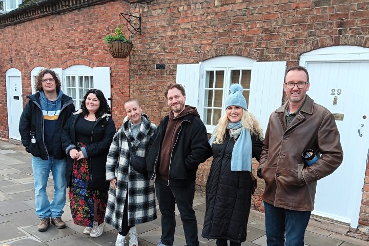 Group of six people standing on a sidewalk in front of a brick building with white doors.