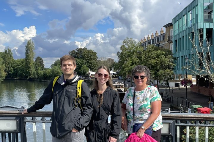 Three people standing on a riverside walkway with cloudy sky in the background.