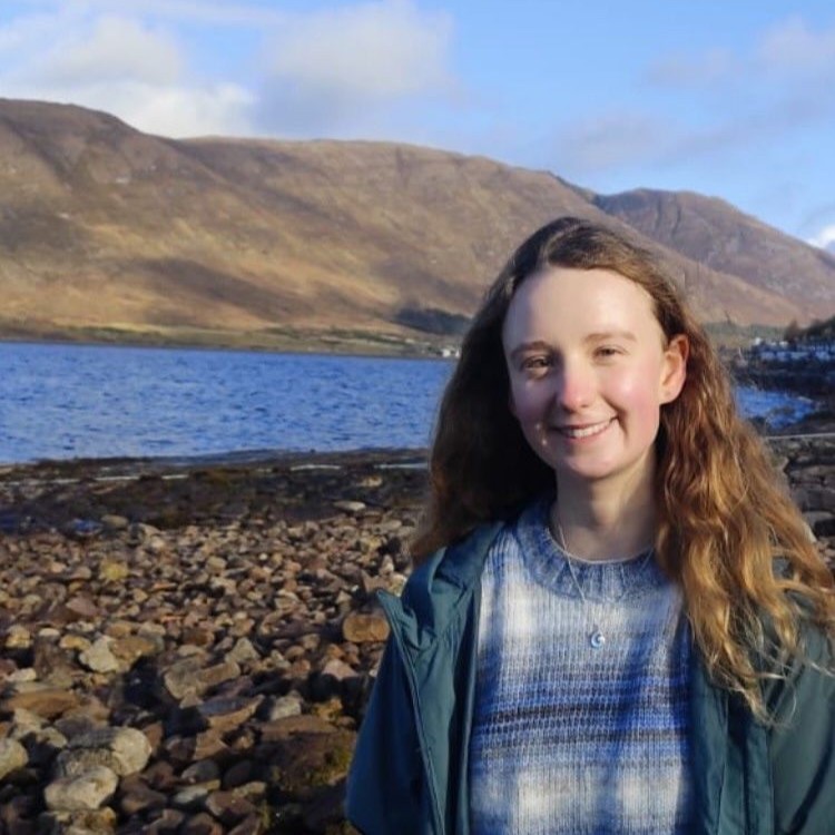 Person with long hair smiling by a rocky lakeside with mountains in the background.