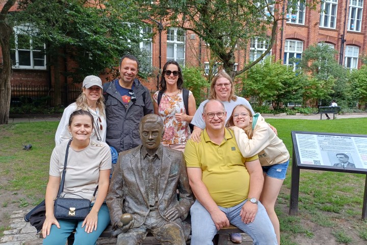 Group of people posing with a statue outdoors near a plaque and red brick building.