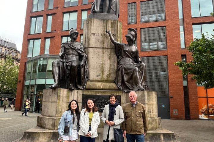 Four people stand in front of a statue labeled 'Wellington' with a brick building in the background.