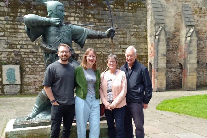 Four people stand smiling in front of a large statue of an archer with a bow, on a stone-paved area.