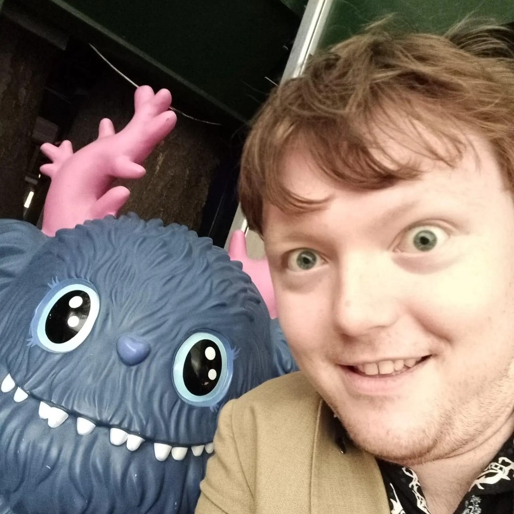 Person taking a selfie with a blue furry monster statue with pink antlers.