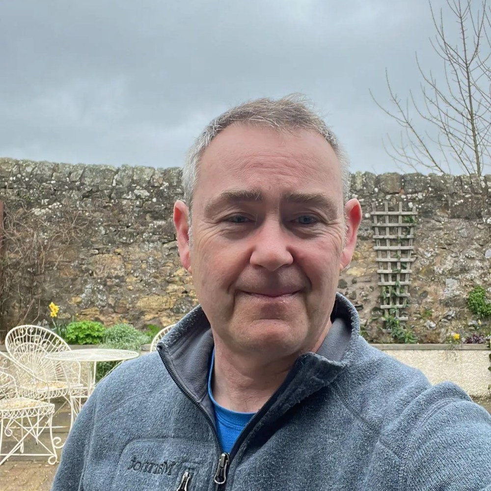Person in a gray jacket smiling in a garden with stone wall and outdoor furniture.