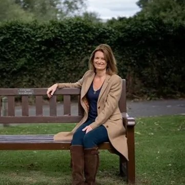 Woman in brown coat sitting on a bench in a park with greenery in the background.