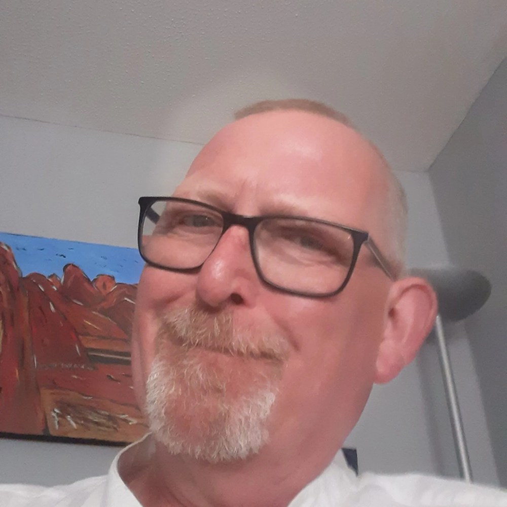 Smiling man with glasses and goatee in front of a landscape painting.
