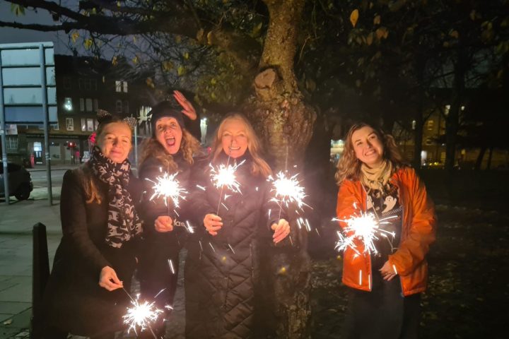 Four people holding sparklers in a park at night, standing under a tree.
