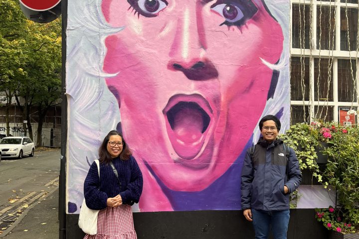 Two people pose in front of a colorful mural of a surprised face in an urban area.