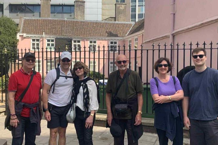 Six people posing in front of a pink building and metal fence on a sunny day.