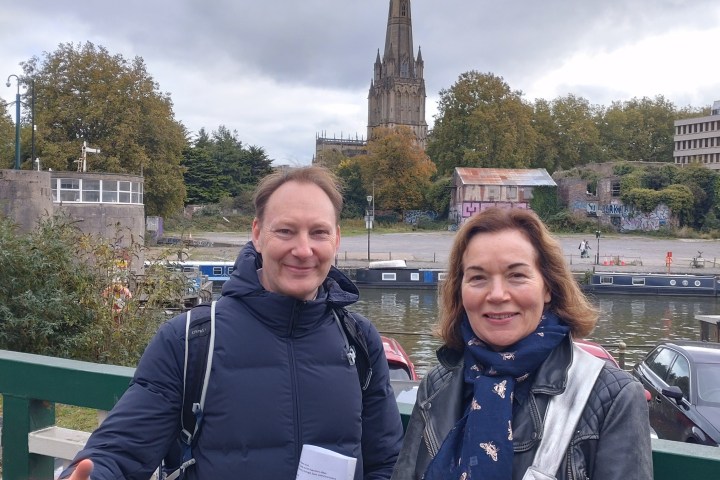 Two people smiling on a bridge with a historic church and trees in the background.