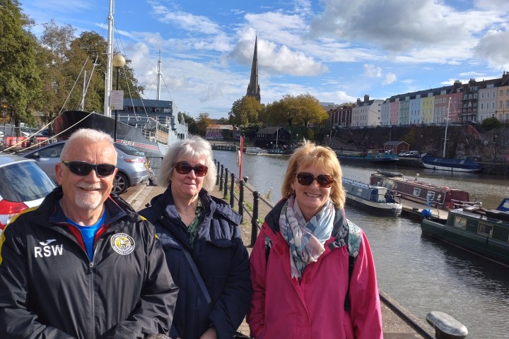 Three people in jackets and sunglasses by a canal with colorful houses and boats in the background.