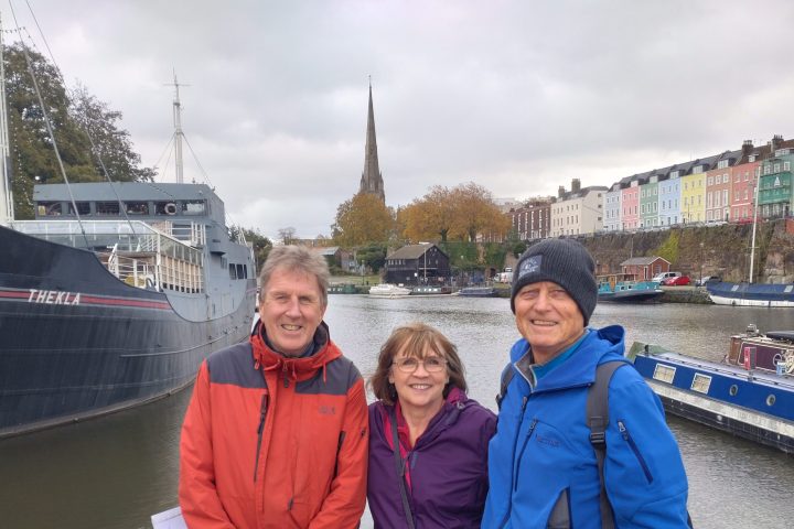 Three people in jackets stand by a waterfront with boats and colorful buildings in the background.