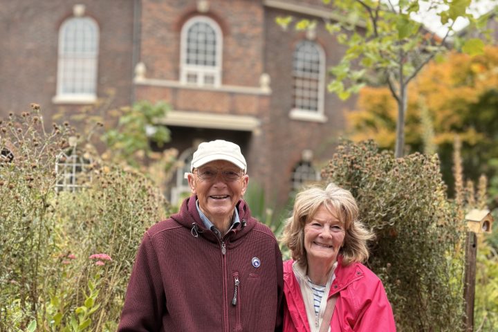 Elderly couple in jackets smiling by garden with brick building in background.