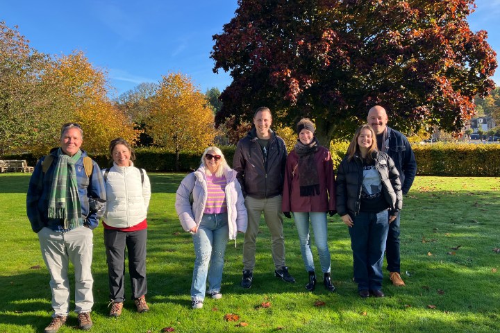 Group of seven people standing on grass in front of a large tree with autumn leaves.