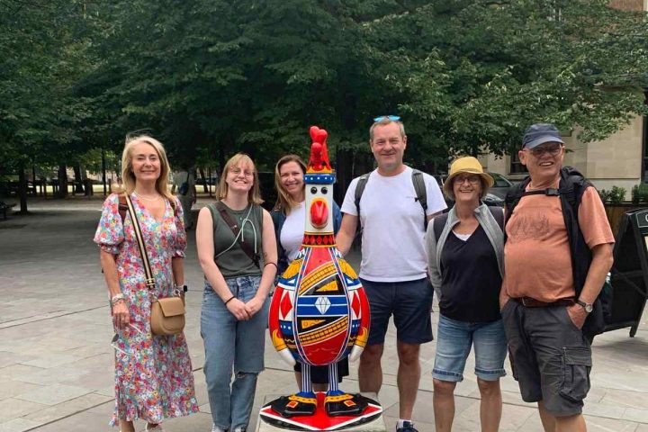Six people stand around a colorful penguin statue outdoors with trees in the background.
