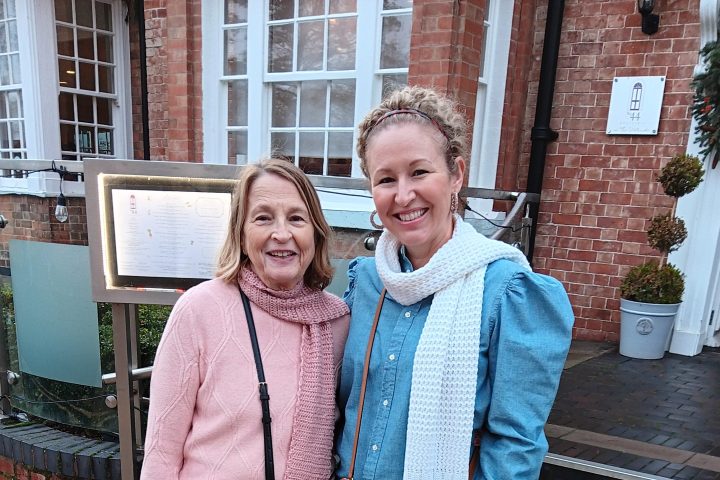 Two smiling women stand outside a building with red bricks and white windows.