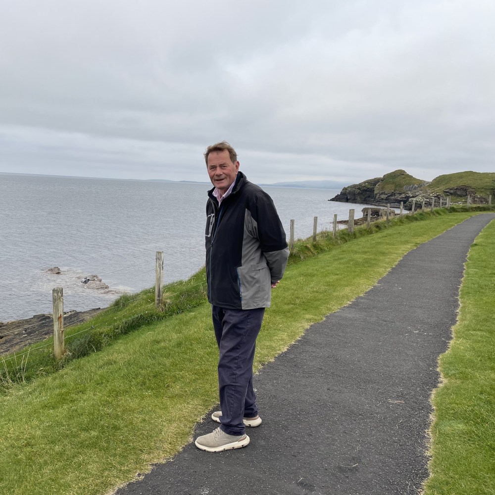 Man on coastal path, wearing a jacket, standing next to grassy area with ocean view.