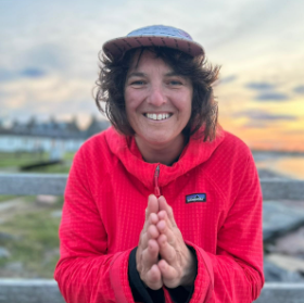 Person in red jacket and cap smiling with hands together, sunset background.