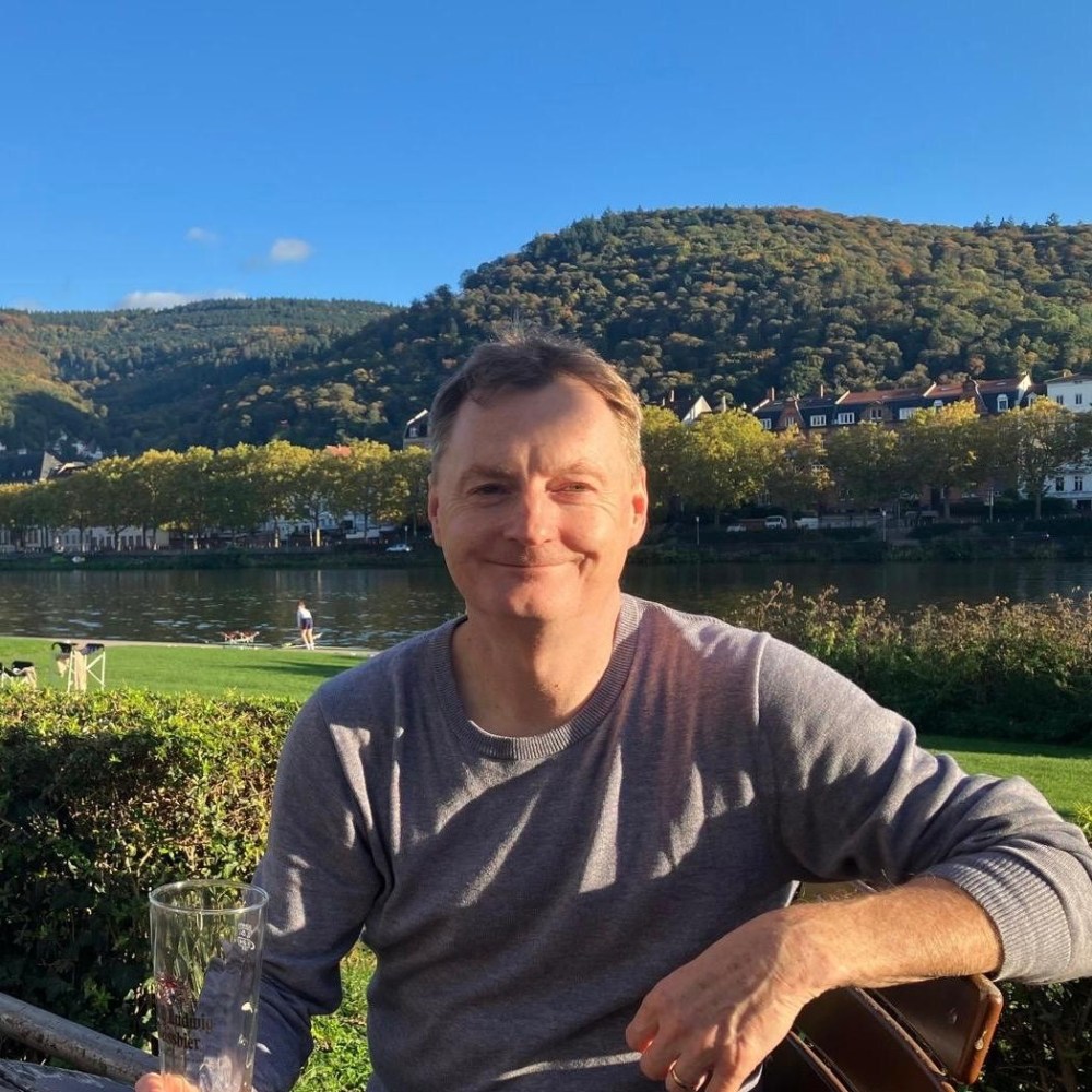 Smiling man in gray sweater sits outdoors by a river with trees and hills in the background.