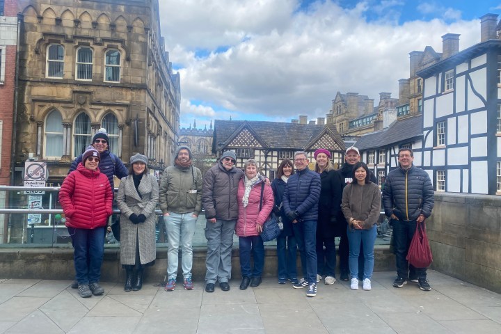 Group of twelve people bundled in jackets standing in front of historic buildings under a cloudy sky.