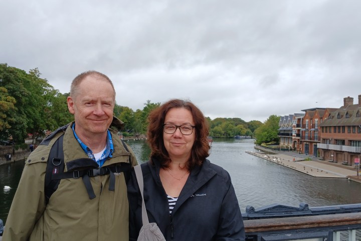 Two people standing on a bridge over a river with buildings and trees in the background.