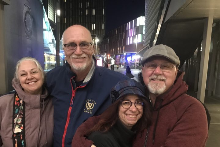Two smiling couples standing together outside at night with city lights in the background.