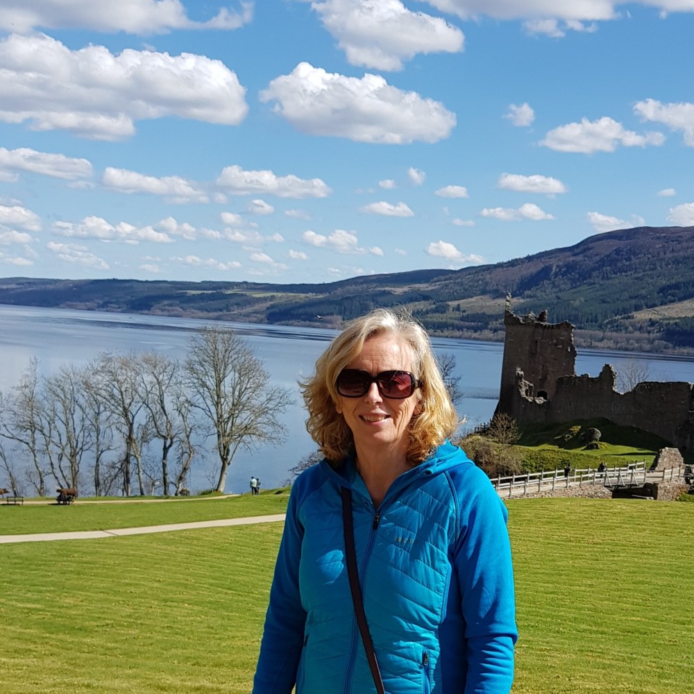 Woman in blue jacket stands in front of lake and castle ruins under a partly cloudy sky.