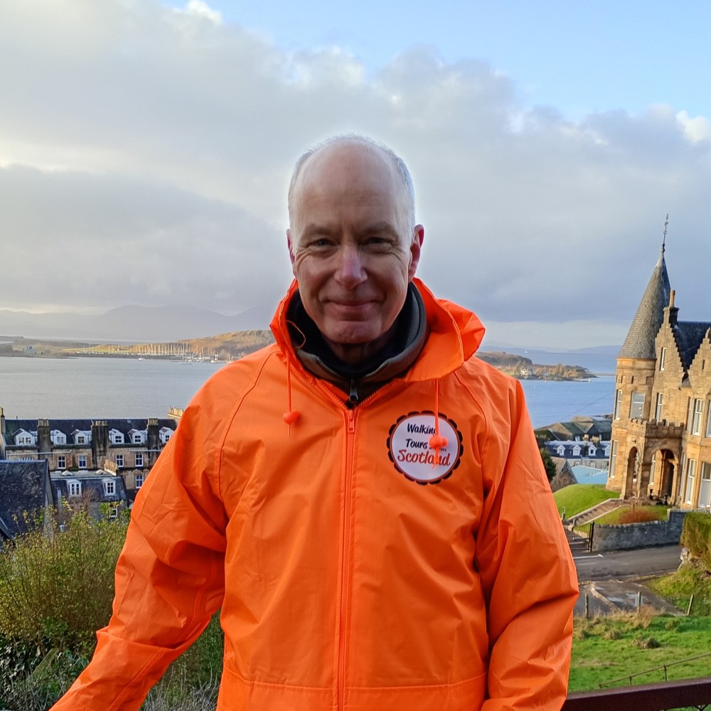 Man in orange jacket poses with scenic coastal town and hills in the background on a cloudy day.