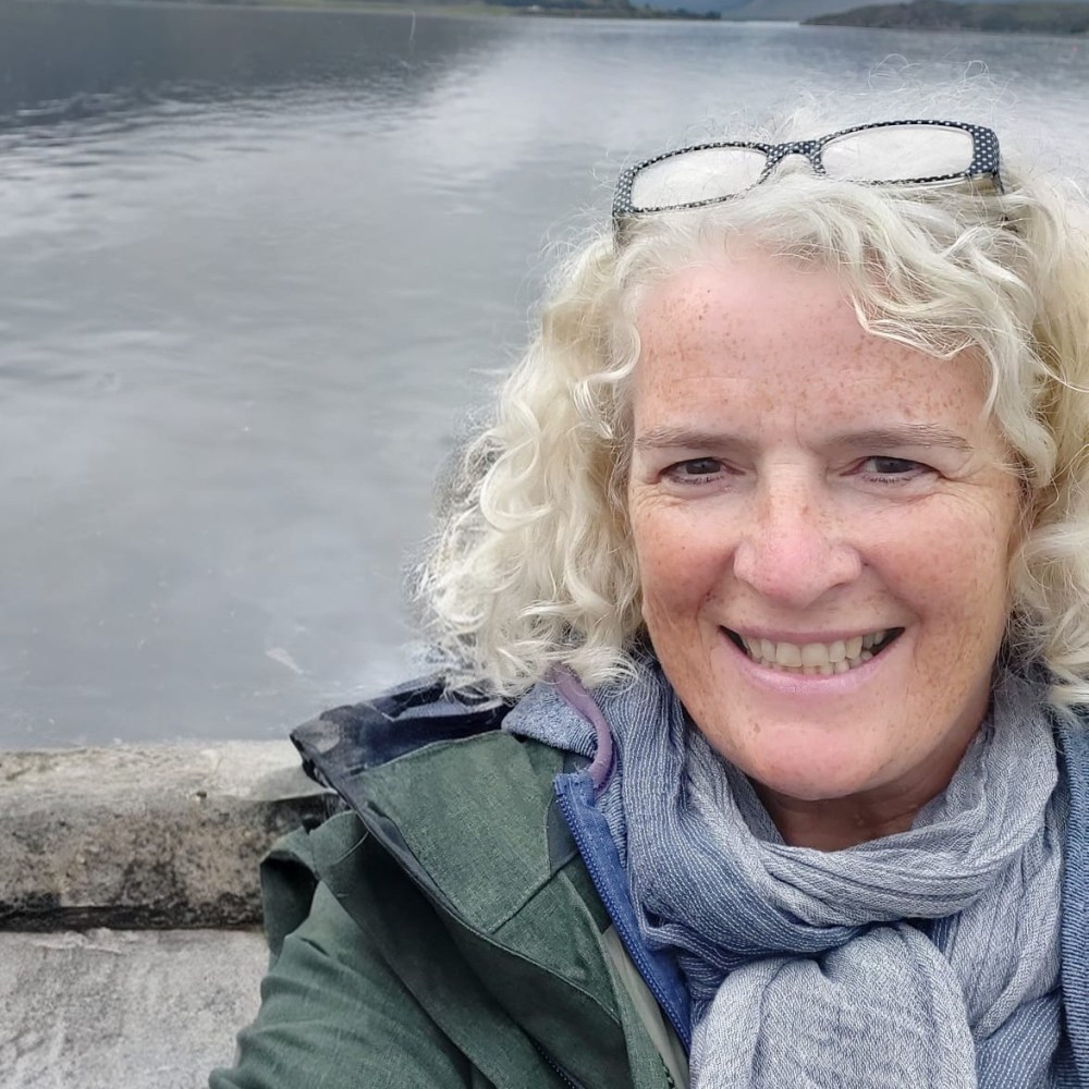Smiling person with curly hair, standing by a lake, wearing glasses and a scarf.