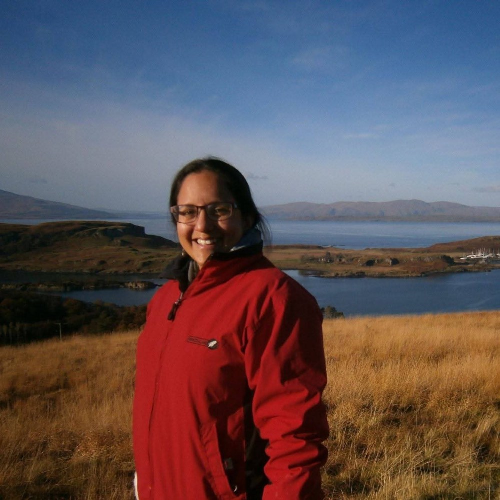 Person in red jacket smiling in a grassy field with a lake and mountains in the background.