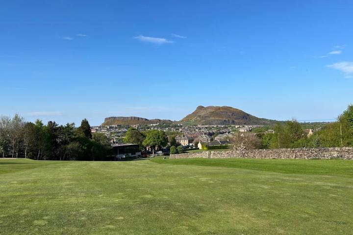 Green field with distant hills and blue sky, with a village and trees in the background.