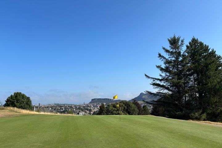 Golf course with flag, trees, and distant hills under a clear blue sky.
