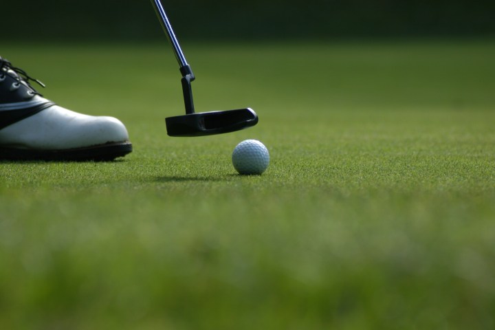 Golf ball near putter on green with golfer’s shoes visible.