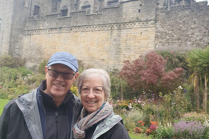 Two people smiling in front of an old stone building with a garden.
