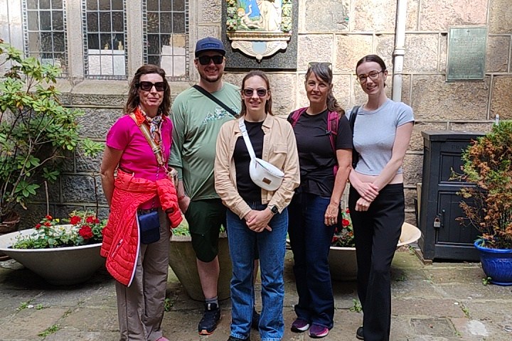 Group of five people posing outdoors by a stone building with a religious plaque.