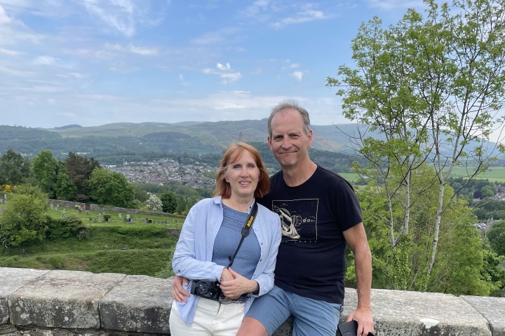 Couple posing by a stone wall with a scenic view of hills and trees in the background.