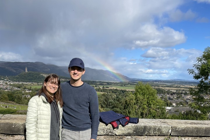 Two people stand by a stone wall with a scenic view and a rainbow in the background.