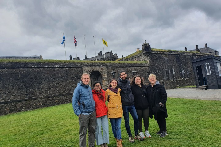 Group of people standing on grass in front of a stone fort under cloudy skies.