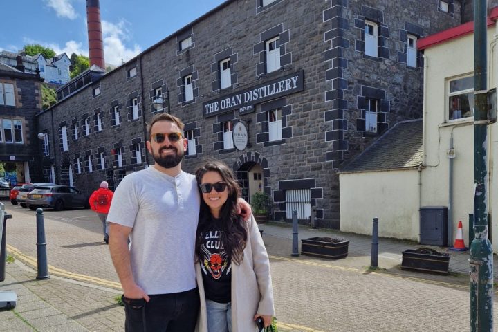 Two people and a dog in front of The Oban Distillery on a sunny day.
