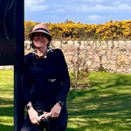 Person in hat smiling by a black post, with greenery and stone wall in background.