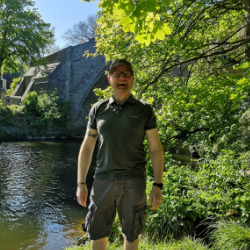 Person standing by a river, surrounded by lush greenery.