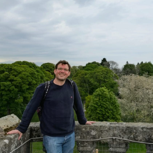 Person smiling at a stone railing with lush trees in the background.