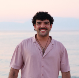 Smiling person in pink shirt stands by the sea at sunset.