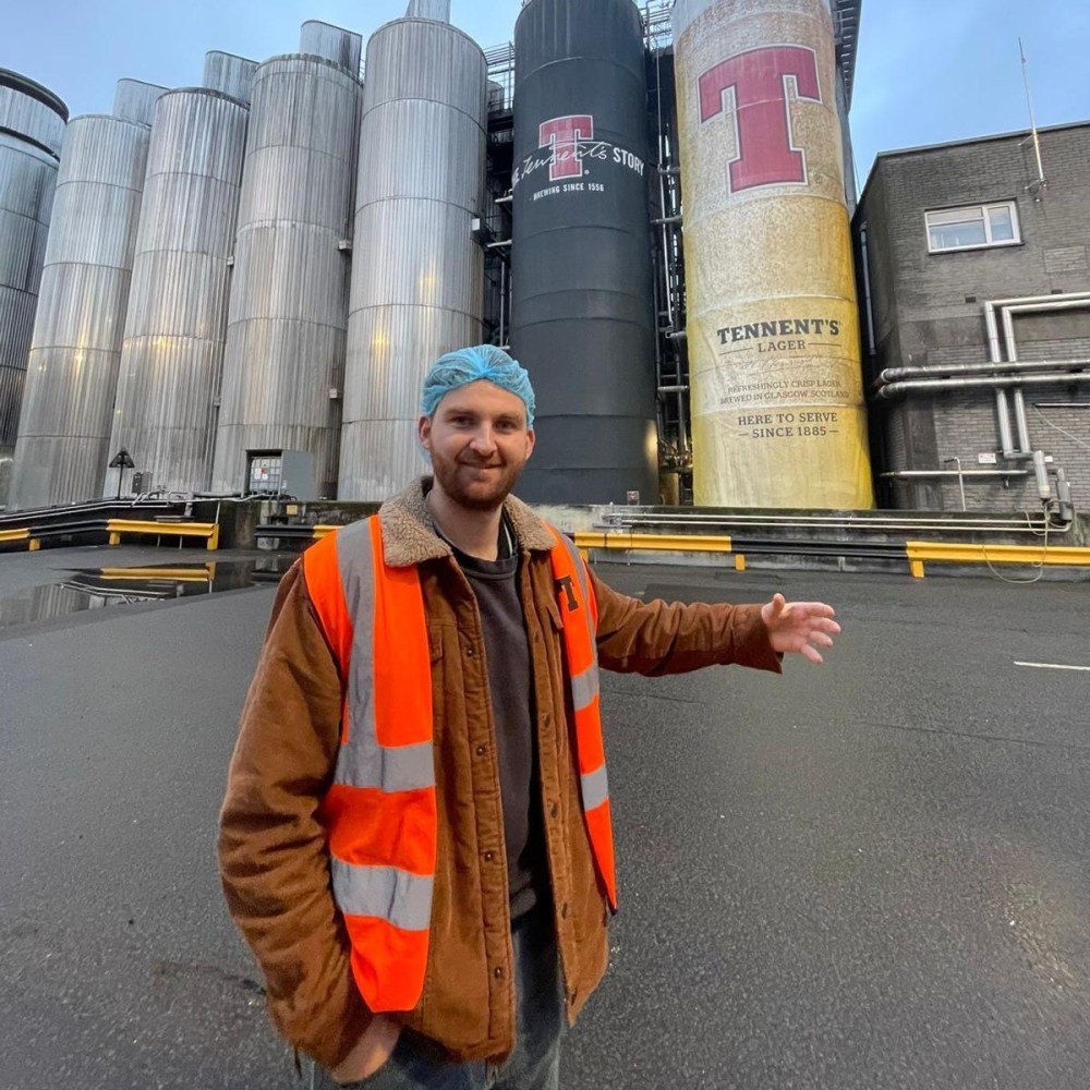 Man in orange vest and blue cap in front of large brewery tanks.