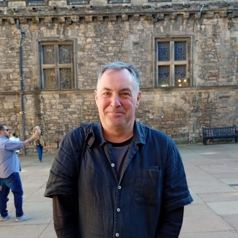Man in dark shirt stands smiling in front of stone building with windows.